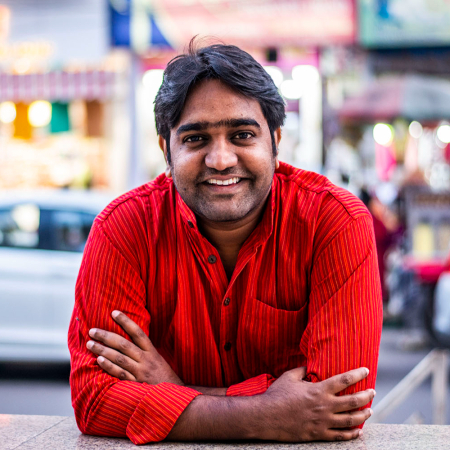 Portrait of Rafiul Alom Rahman. A person with black hair and a three-day beard wears a red shirt and sits casually in an urban environment.