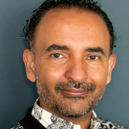 Portrait of Rafiul Alom Rahman. A person with black hair and a three-day beard wears a red shirt and sits casually in an urban environment.