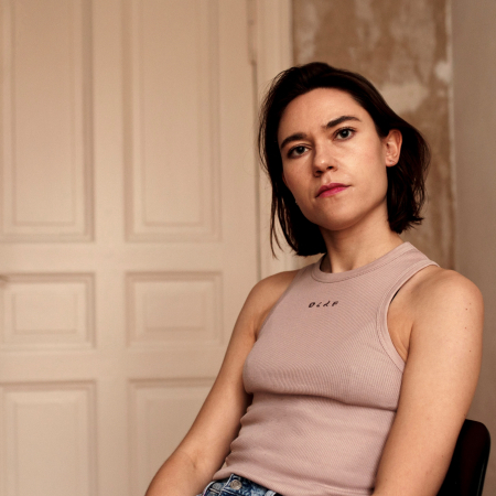 Paulina Lorenz in portrait. A person with shoulder-length hair sits casually on a chair in an old flat.