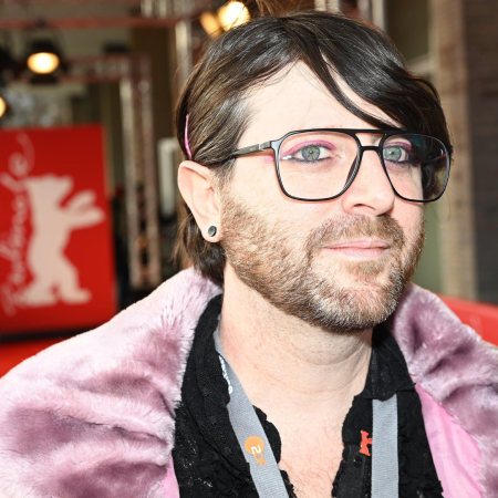 A portrait of Bartholomew Sammut. A person with a short full beard and black glasses wears pink eye shadow with black eyeliner and a fluffy, dusky pink coat. The medium-length brown fringe is loosely tucked behind the left ear.