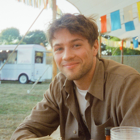 Connor Jessup im Portrait. Eine Person mit dunkelblonden 3-Tage-Bart und gestyltem Fassonschnitt sitzt auf einer Bank auf einem Festival.