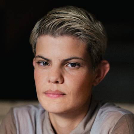 A person with short, blonde streaked hair and brown eyes wears an iridescent blouse.